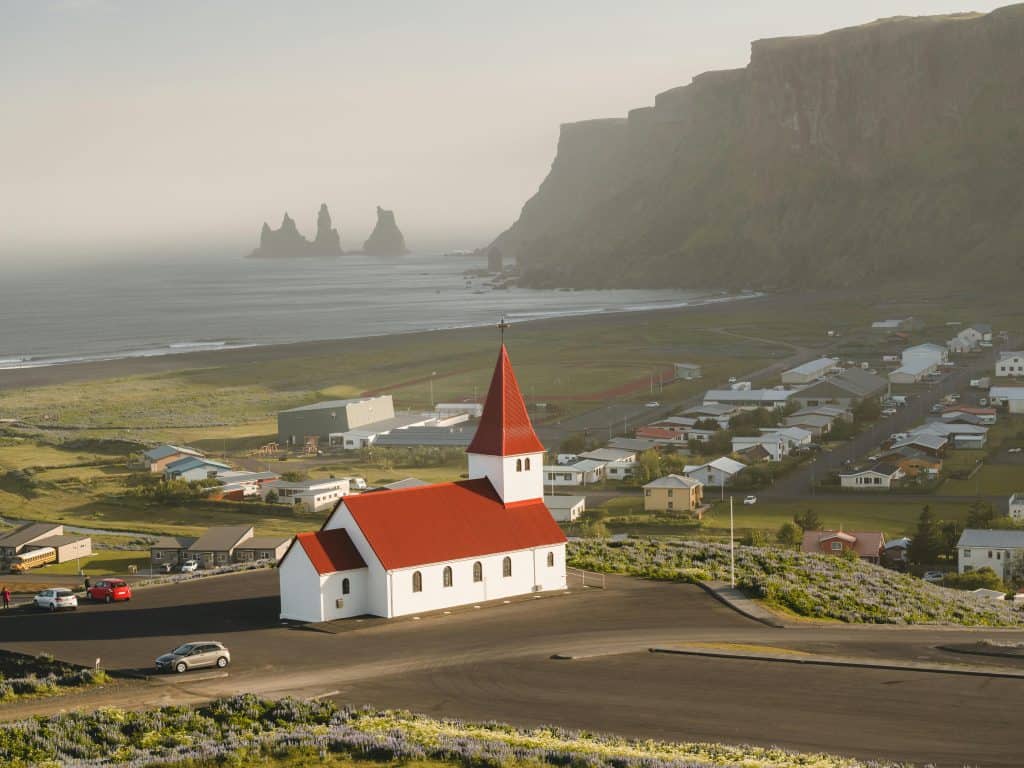 Aerial view of the iconic red-roofed Vik Church with Reynisdrangar sea stacks along the stunning Icelandic coastline.